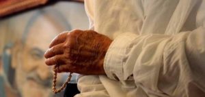 Ram Dass's hand holding a mala, with photo of his guru Maharajji softly in the background.